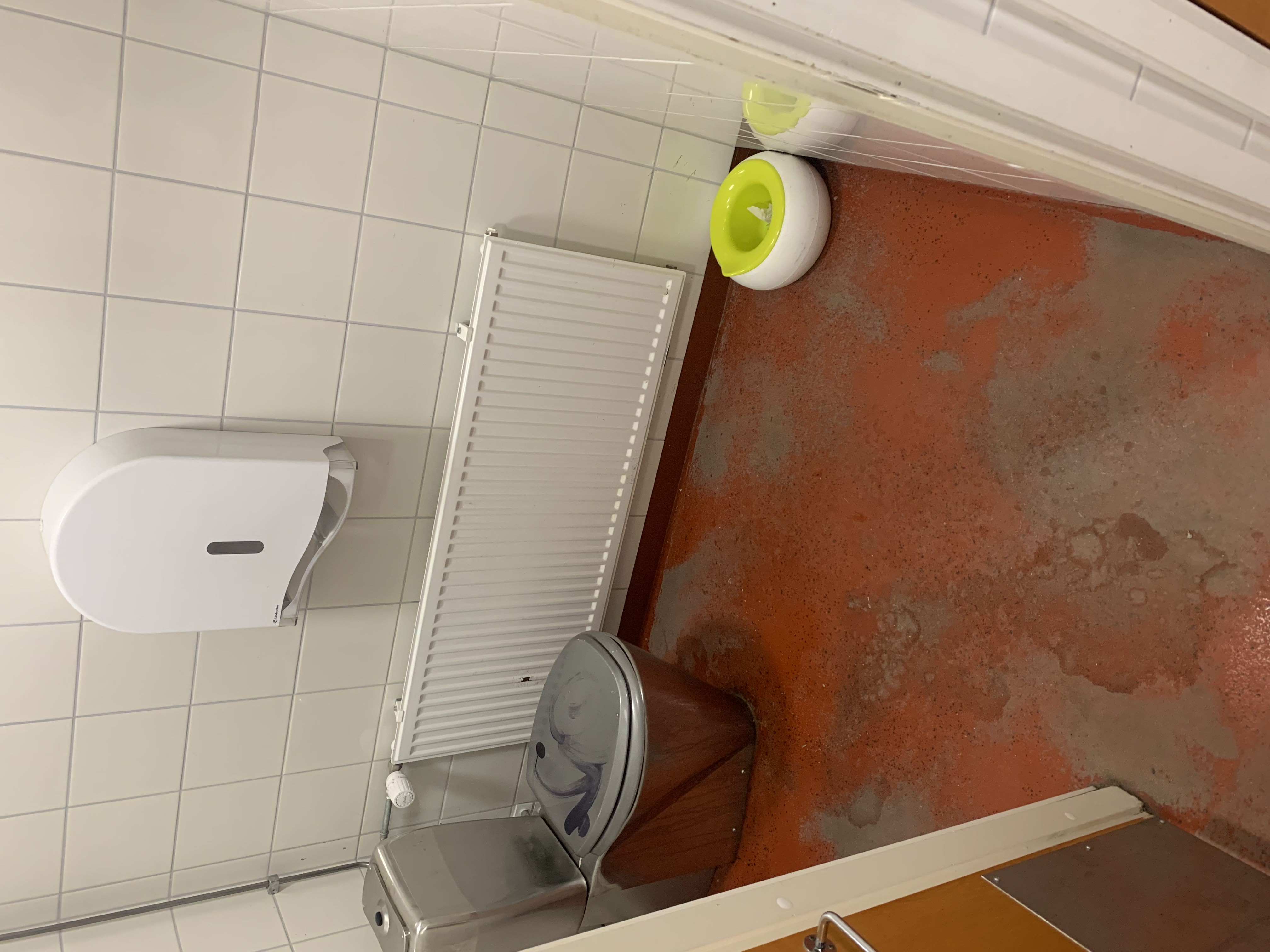 Kuvassa WC-koppi sisältä, metallinen pönttö vasemmalla ja potta lattialla oikeassa kulmassa.