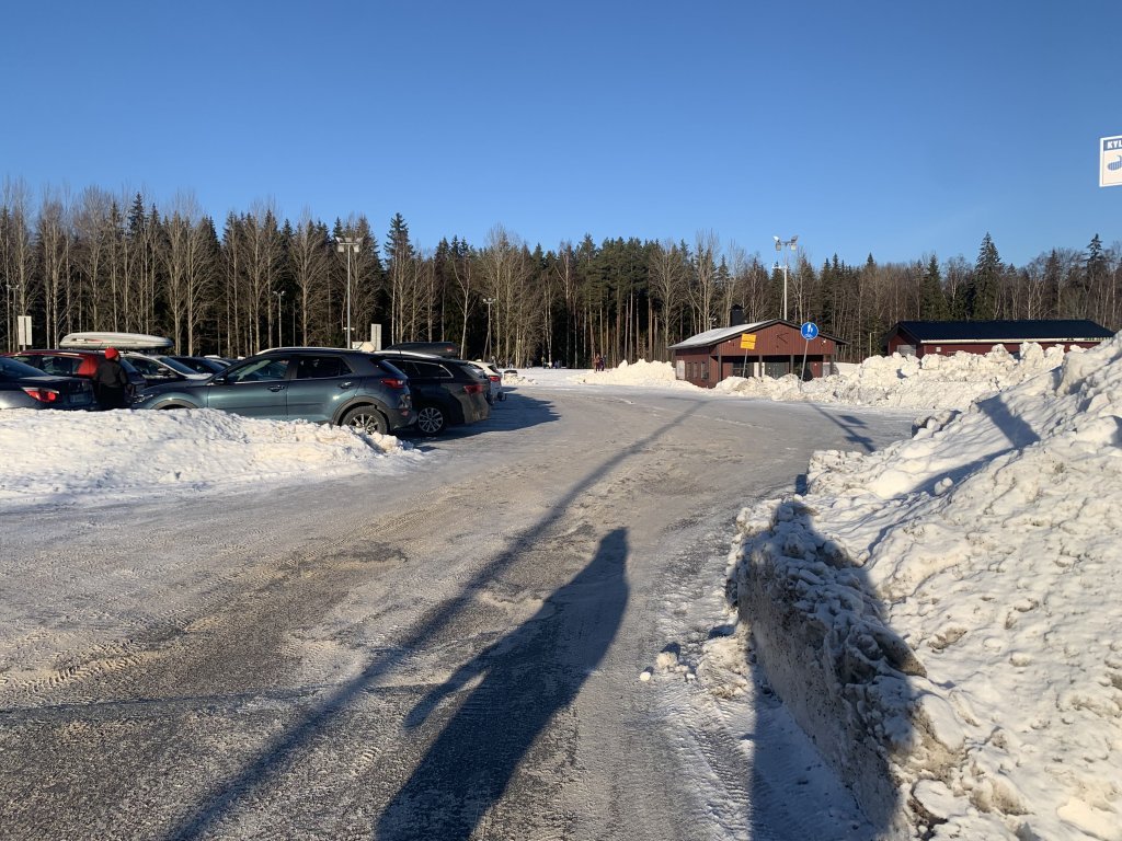 Kuvassa paljon autoja parkkipaikalla harjakattoisen rakennuksen näkyessä parkkipaikan oikeassa kulmassa. Lumisen kentän takana näkyy metsää.